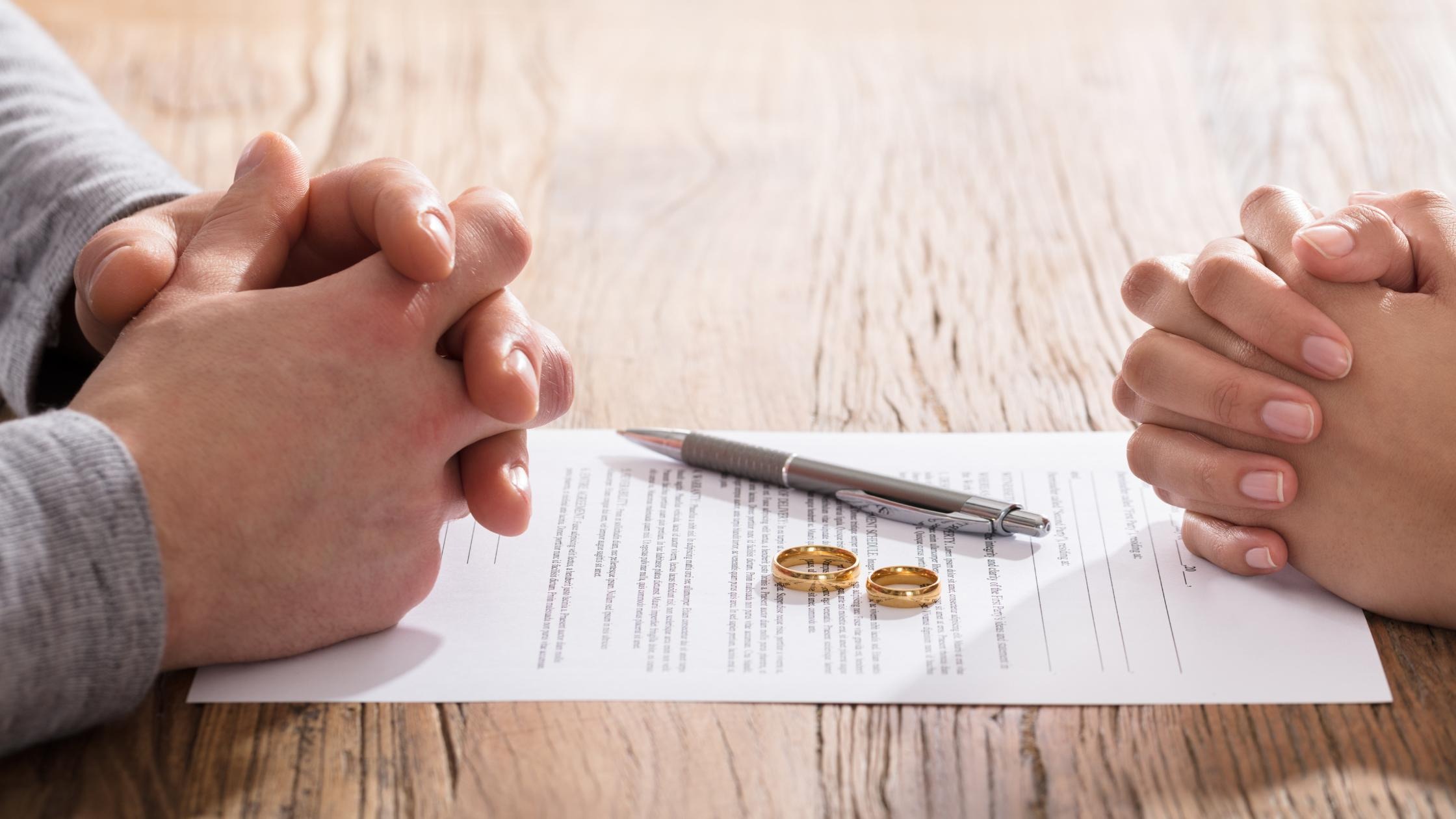 Two hands clasped across a divorce agreement document with wedding rings and a pen placed on the table.