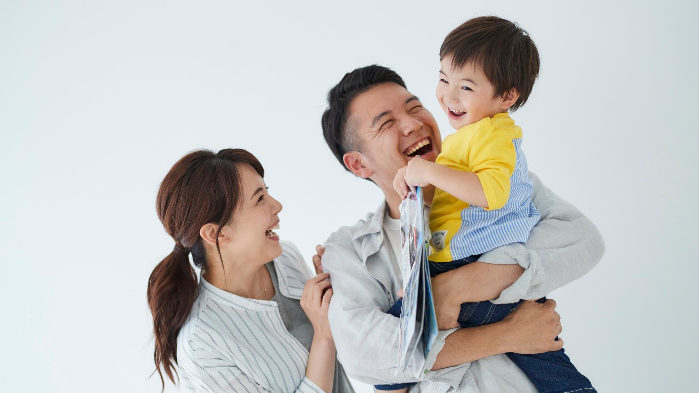 Happy family with parents laughing together with their young child in a yellow shirt against a white background.