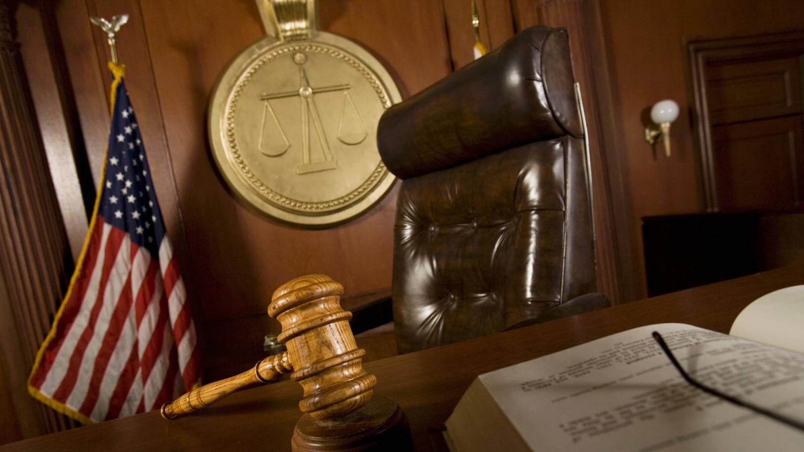 Judge’s bench in a courtroom with a gavel, legal documents, the U.S. flag, and the scales of justice emblem in the background.